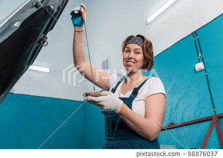 Portrait of young woman mechanic in overall and gloves repairs oil dipstick of car. Bottom view. The concept of women's equality Portrait of young woman mechanic in overall and gloves repairs oil dipstick of car. Bottom view. The concept of women's equality 88876037