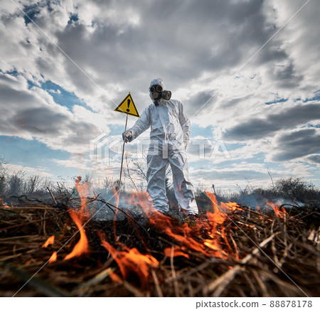 Firefighter ecologist extinguishing fire in field. Man in protective radiation suit and gas mask near burning grass with smoke, holding warning sign with exclamation mark. Natural disaster concept. 88878178