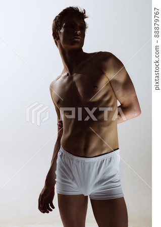 Close-up portrait of young handsome shirtless muscled man wearing white boxer-briefs standing isolated on gray background. Natural beauty of male body Close-up portrait of young handsome shirtless muscled man wearing white boxer-briefs standing isolated on gray background. Natural beauty of male body 88879767