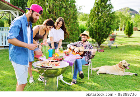 a group of friends in the park at a picnic are barbecued, chatting, smiling and playing with a Labrador Retriever dog against the backdrop of greenery and a gazebo 88889736
