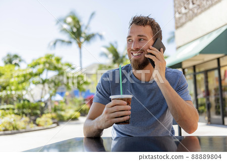 Man calling using phone making a call drinking cold coffee drink sitting at cafe terrace holding smartphone talking on mobile smart phone in summer. Handsome young casual man smiling happy outside 88889804