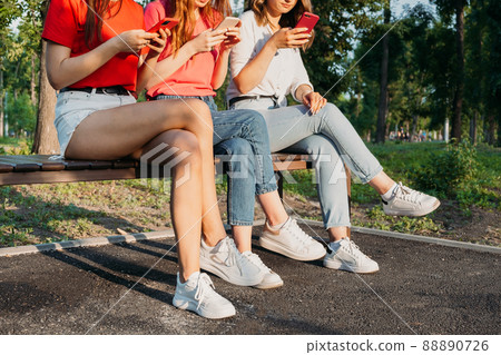 Diverse three girl friends using their phones outdoors. Group gen z young people using mobile smartphone sitting on bench in summer park 88890726