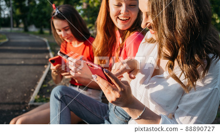 Diverse three girl friends using their phones outdoors. Group gen z young people using mobile smartphone sitting on bench in summer park Diverse three girl friends using their phones outdoors. Group gen z young people using mobile smartphone sitting on bench in summer park 88890727