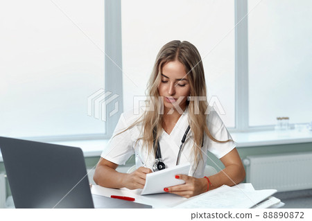 female doctor using laptop and writing notes in medical journal sitting at desk. 88890872