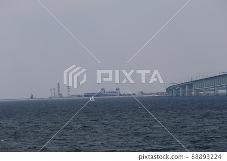 A view of an artificial island with a hazy blue sky, a blue sea surface, a yacht, a bridge, a building, and an airport. 88893224