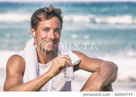 Healthy fit young man athlete drinking water bottle after run workout on beach listening to music phone app with earphones. Smiling portrait Healthy fit young man athlete drinking water bottle after run workout on beach listening to music phone app with earphones. Smiling portrait 88894747