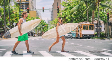 Surfers going surfing in Waikiki beach, Honolulu, Hawaii. USA summer travel vacation happy couple friends walking crossing city street carrying surfboards. Asian woman, Caucasian young man Surfers going surfing in Waikiki beach, Honolulu, Hawaii. USA summer travel vacation happy couple friends walking crossing city street carrying surfboards. Asian woman, Caucasian young man 88898761