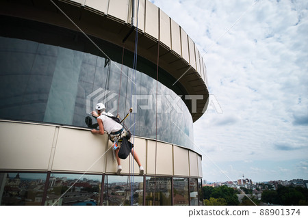 Industrial mountaineering worker professional cleaner hanging on climbing rope and cleaning window of high-rise building. Man window washer washing skyscraper facade. Industrial mountaineering worker professional cleaner hanging on climbing rope and cleaning window of high-rise building. Man window washer washing skyscraper facade. 88901374