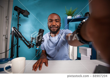 Wide angle view of famous vlogger taking a selfie in vlog studio smiling at camera in front of recording microphone. Pov shot of social media content creator at desk with live broadcasting setup. Wide angle view of famous vlogger taking a selfie in vlog studio smiling at camera in front of recording microphone. Pov shot of social media content creator at desk with live broadcasting setup. 88907204