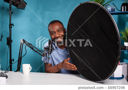 Portrait of photography equipment reviewer presenting beauty dish honeycomb grid in vlogging studio. Vlogger explaining features of studio flash light modifier sitting at desk with microphone. Portrait of photography equipment reviewer presenting beauty dish honeycomb grid in vlogging studio. Vlogger explaining features of studio flash light modifier sitting at desk with microphone. 88907206