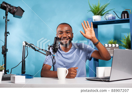 Happy vlogger waving hello at audience during online live show sitting at desk with laptop and broadcasting setup. Content creator doing greeting hand gesture in recording studio with professional Happy vlogger waving hello at audience during online live show sitting at desk with laptop and broadcasting setup. Content creator doing greeting hand gesture in recording studio with professional 88907207