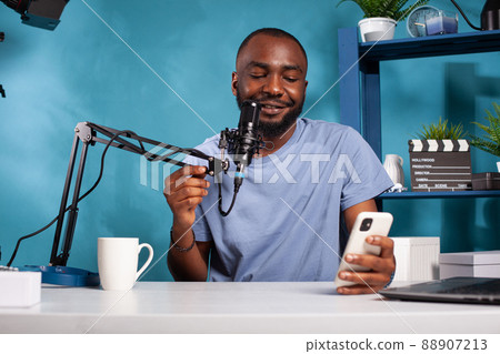 Influencer having questions and answers session using microphone in recording desk in vlogging studio. African american vlogger reading live text messages from fans looking at smartphone screen. Influencer having questions and answers session using microphone in recording desk in vlogging studio. African american vlogger reading live text messages from fans looking at smartphone screen. 88907213
