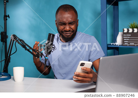 Portrait of influencer crying after reading on smartphone insulting comments from online social media account . Vlogger sitting at desk in vlogging studio deeply hurt by online bully. Portrait of influencer crying after reading on smartphone insulting comments from online social media account . Vlogger sitting at desk in vlogging studio deeply hurt by online bully. 88907246