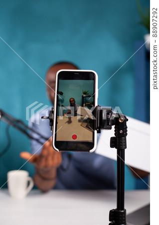Selective focus on mobile phone recording influencer hosting online giveaway holding product box in vlogging studio. Closeup of live vlog setup with smarthone on stand filming smiling vlogger. 88907292