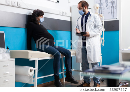 Pregnant adult talking about childbirth with health physician during covid 19 pandemic. General practitioner doing consultation with woman expecting child at checkup visit in medical cabinet. 88907337