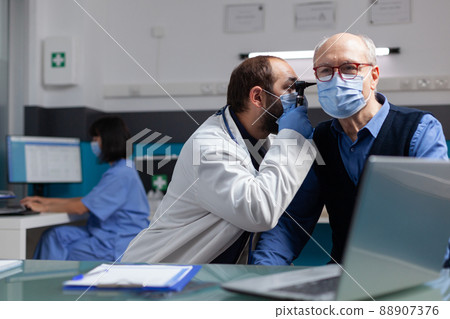 Specialist using otoscope to do ear examination on elder man, doing otolaryngology consultation with patient. Doctor with face mask examining infection to give prescription treatment. 88907376