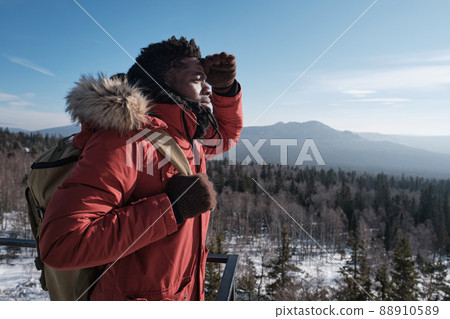 Young African American man wearing backpack standing at viewing platform on mountain top on winter day looking into distance Young African American man wearing backpack standing at viewing platform on mountain top on winter day looking into distance 88910589