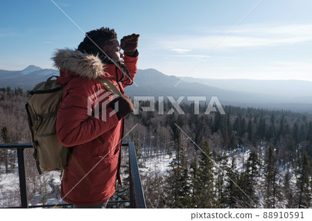Young African American traveler wearing backpack standing at observation deck on mountain top on winter day looking into distance Young African American traveler wearing backpack standing at observation deck on mountain top on winter day looking into distance 88910591