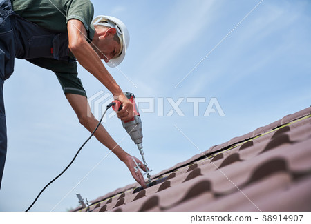 Worker prepearing to installation photovoltaic solar panel system on the roof of house. Man installer working with electric screwdriver, wearing uniform and helmet, on blue sky background. 88914907