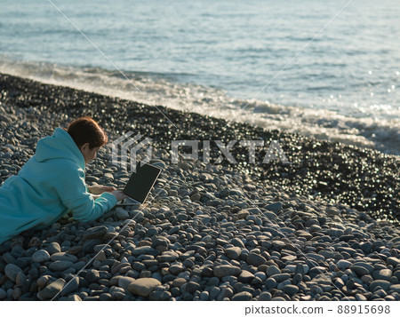 Caucasian woman lying on her stomach on a pebble beach and typing on a laptop. Remote work freelancer. Caucasian woman lying on her stomach on a pebble beach and typing on a laptop. Remote work freelancer. 88915698