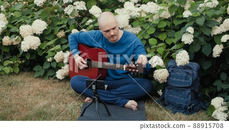 Man with an acoustic guitar is sitting in a park in nature and tuning the instrument using an app on his phone. The concept of travel, hiking and hobbies Man with an acoustic guitar is sitting in a park in nature and tuning the instrument using an app on his phone. The concept of travel, hiking and hobbies 88915708