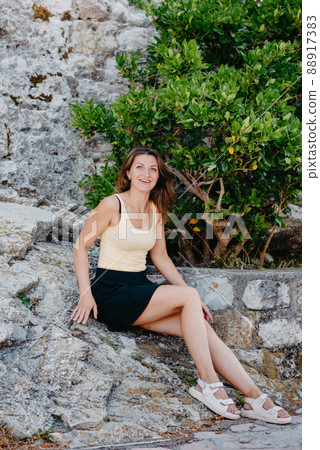 Beautiful girl sits on the stone wall of the old town of Budva . The girl sits on a stone parapet, Budva, Montenegro Beautiful girl sits on the stone wall of the old town of Budva . The girl sits on a stone parapet, Budva, Montenegro 88917383