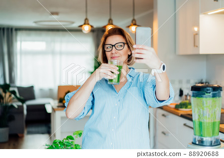Middle aged woman making selfie with just made glass of detox shake, green smoothie in the kitchen. Healthy dieting, eating, cooking, weight loss program. Blogger creating content for social media. 88920688