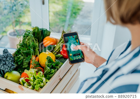 Top view woman holding wooden box with fresh vegetables and phone with active online mobile application with Veganuary diet program. New Start of a healthy lifestyle, weight loss concept. Top view woman holding wooden box with fresh vegetables and phone with active online mobile application with Veganuary diet program. New Start of a healthy lifestyle, weight loss concept. 88920715