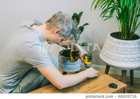 A young man with an electrical screwdriver assembles a tv stand console according to instructions in his new house. Man assembling furniture at home using a cordless screwdriver. Selective focus. A young man with an electrical screwdriver assembles a tv stand console according to instructions in his new house. Man assembling furniture at home using a cordless screwdriver. Selective focus. 88920716