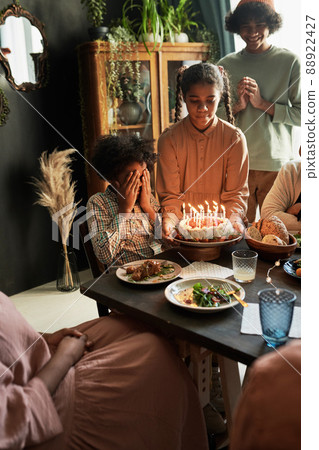African little boy sitting at table with his eyes closed while his family congratulating him with birthday cake African little boy sitting at table with his eyes closed while his family congratulating him with birthday cake 88922427