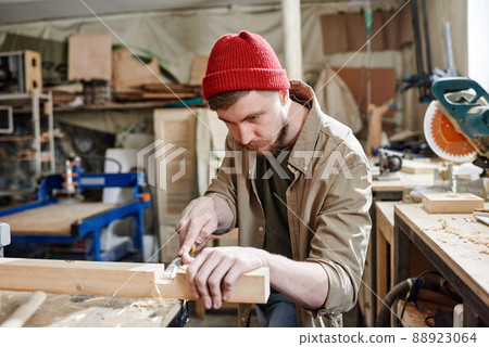 Bearded Caucasian carpenter wearing red knit cap cutting mortise in wood plank using chisel 88923064