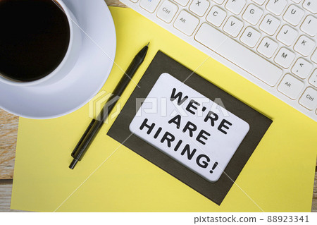 We're are hiring text on white note with yellow sheet on wooden desk. 88923341