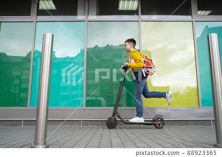 Modern teenager with backpack rides on electric scooter on brick wall background. Boy comes back from school 88923365