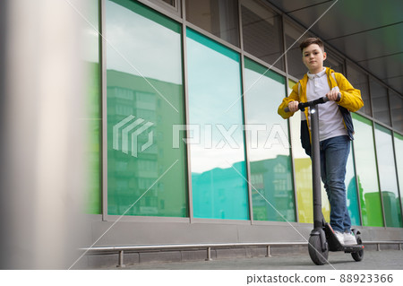 Handsome teenager with backpack rides on electric scooter down the city street Handsome teenager with backpack rides on electric scooter down the city street 88923366