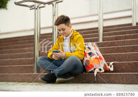 Schoolboy teenager sits on the stairs with phone in his hands. Concept of modern generation addicted to gadgets Schoolboy teenager sits on the stairs with phone in his hands. Concept of modern generation addicted to gadgets 88923780