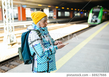 Old happy woman with backpack in yellow hat waiting train on station platform and using smart phone on urban background. Railroad transport concept Old happy woman with backpack in yellow hat waiting train on station platform and using smart phone on urban background. Railroad transport concept 88927666