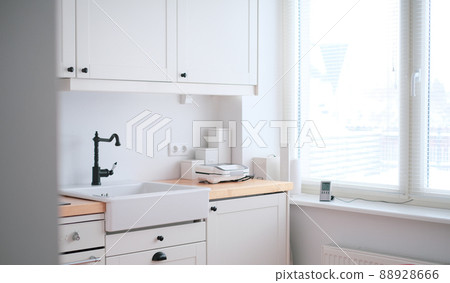 The interior of a kitchen in white tones with a sink, sandwich maker, stove, oven, dining table and a light window 88928666