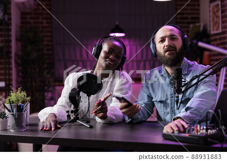Male presenter and guest recording podcast vlog discussion in studio with neon lights. Multiethnic team of people filming video conversation on camera, broadcasting on social media. Male presenter and guest recording podcast vlog discussion in studio with neon lights. Multiethnic team of people filming video conversation on camera, broadcasting on social media. 88931883