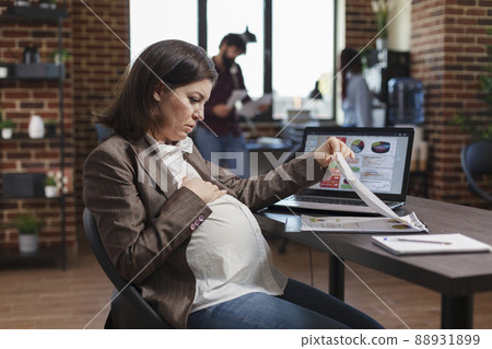 Pregnant woman reviewing agency documentation and business analytics results while sitting at office desk. Marketing company employee analyzing future business plan regarding profits. Pregnant woman reviewing agency documentation and business analytics results while sitting at office desk. Marketing company employee analyzing future business plan regarding profits. 88931899