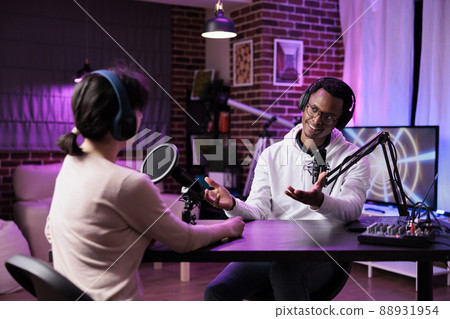 Multiethnic team of people meeting in studio to record podcast show, using camera and sound equipment. Man and woman having fun with chat on live broadcast for social media channel. Multiethnic team of people meeting in studio to record podcast show, using camera and sound equipment. Man and woman having fun with chat on live broadcast for social media channel. 88931954