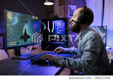 Young adult playing action shooting video games competition in living room with neon lights. Male player streaming live rpg gameplay tournament on computer, enjoying shooter play. Young adult playing action shooting video games competition in living room with neon lights. Male player streaming live rpg gameplay tournament on computer, enjoying shooter play. 88931956
