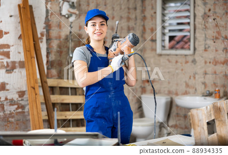 Female builder in blue overalls standing at construction site with chisel hammer Female builder in blue overalls standing at construction site with chisel hammer 88934335