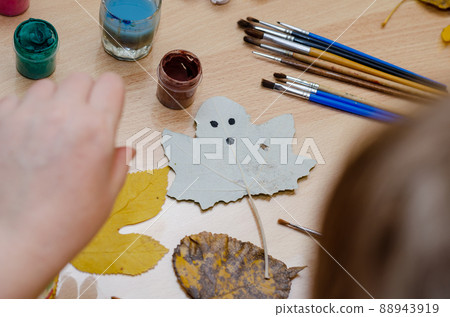 A fallen leaf with a drawing of a ghost's face lies on a beige table. Paints and brushes are nearby. An adult woman's hand reaches for a brush. Inside the room. Selective focus. Series part A fallen leaf with a drawing of a ghost's face lies on a beige table. Paints and brushes are nearby. An adult woman's hand reaches for a brush. Inside the room. Selective focus. Series part 88943919