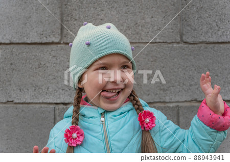 A portrait of a cheerful child against a gray stone wall. A five-year-old girl with her tongue out. A gesticulating daughter with long pigtails. A portrait of a cheerful child against a gray stone wall. A five-year-old girl with her tongue out. A gesticulating daughter with long pigtails. 88943941