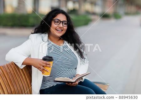 An Indian girl is sitting on a bench, reading a book and drinking coffee. 88945630