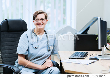 Portrait of happy female doctor in eyeglasses and scrubs sitting in office chair at table with computer Portrait of happy female doctor in eyeglasses and scrubs sitting in office chair at table with computer 88948440