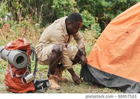 Serious young Afro-American hiker crouching on ground and assembling orange tent in forest 88957583