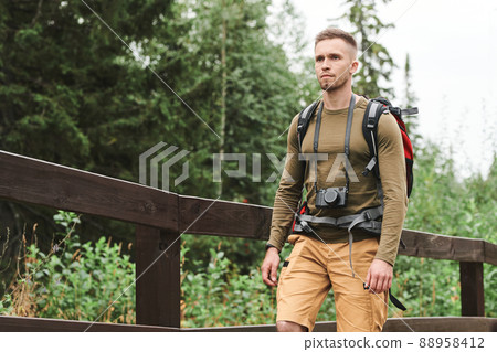 Serious young Caucasian photographer with camera hanging on neck carrying backpack while hiking in forest 88958412