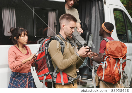 Serious young man looking through photos on camera display while his friends unloading hiking stuff from van Serious young man looking through photos on camera display while his friends unloading hiking stuff from van 88958438