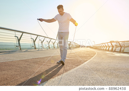 Full length portrait of a muscular build European middle aged man, fit athlete doing jumping exercises, cardio training with skipping rope on the urban city bridge early in the morning on a summer day 88962438